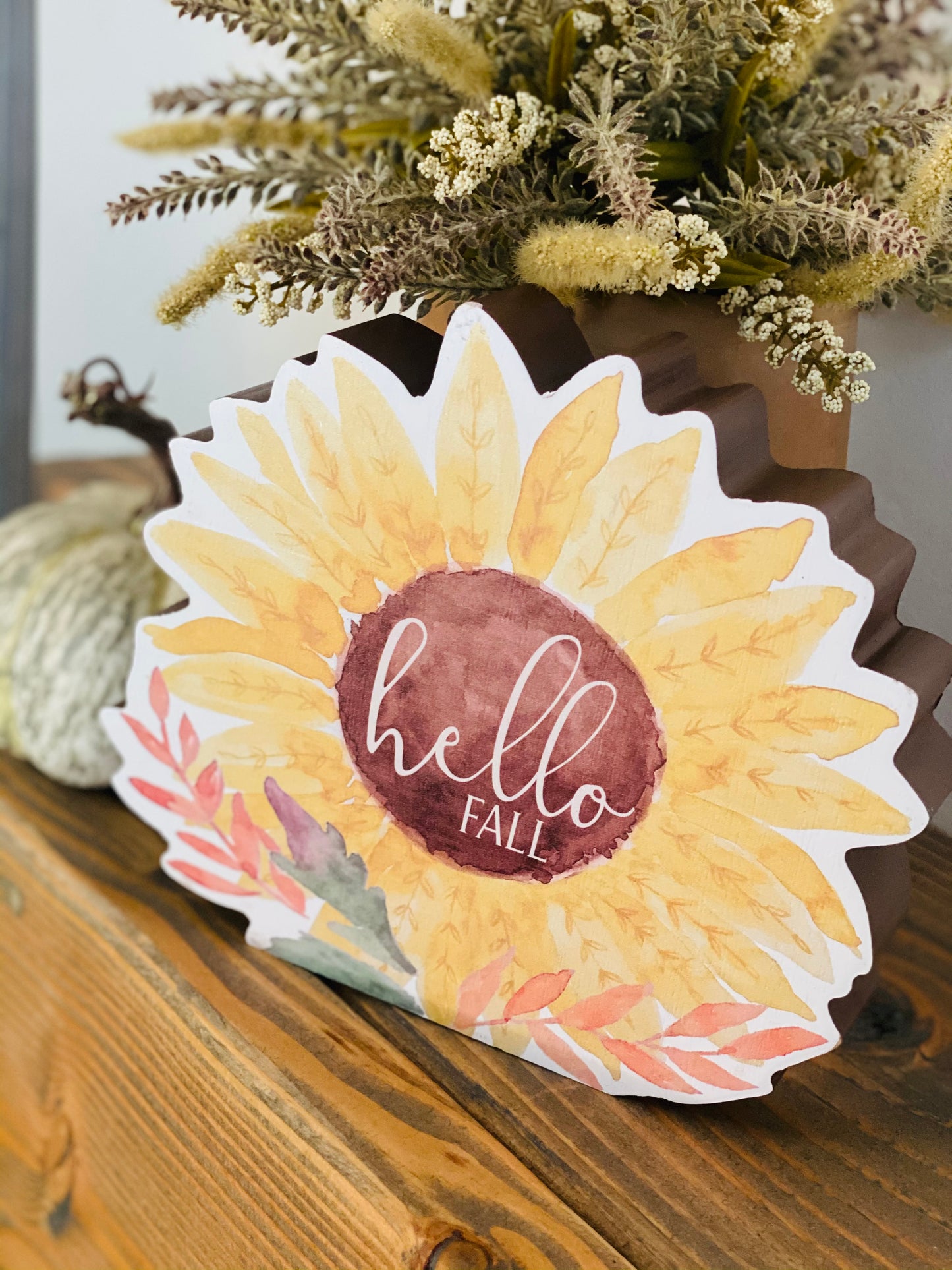 Hello Fall Chunky Watercolor Sunflower Sitter is a freestanding wooden sitter with a sunflower design. Sitter has a watercolor style print with fall-colored leaves at the base and the words, "hello fall" in white text at the center. Sitter is perfect for adding a twist to fall decor 